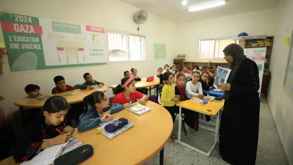 Des enfants assis dans une salle de classe à Gaza, écoutant une enseignante vêtue d'une robe noire. Un panneau en arrière-plan indique '2024 Gaza, L'Éducation Une Urgence'.