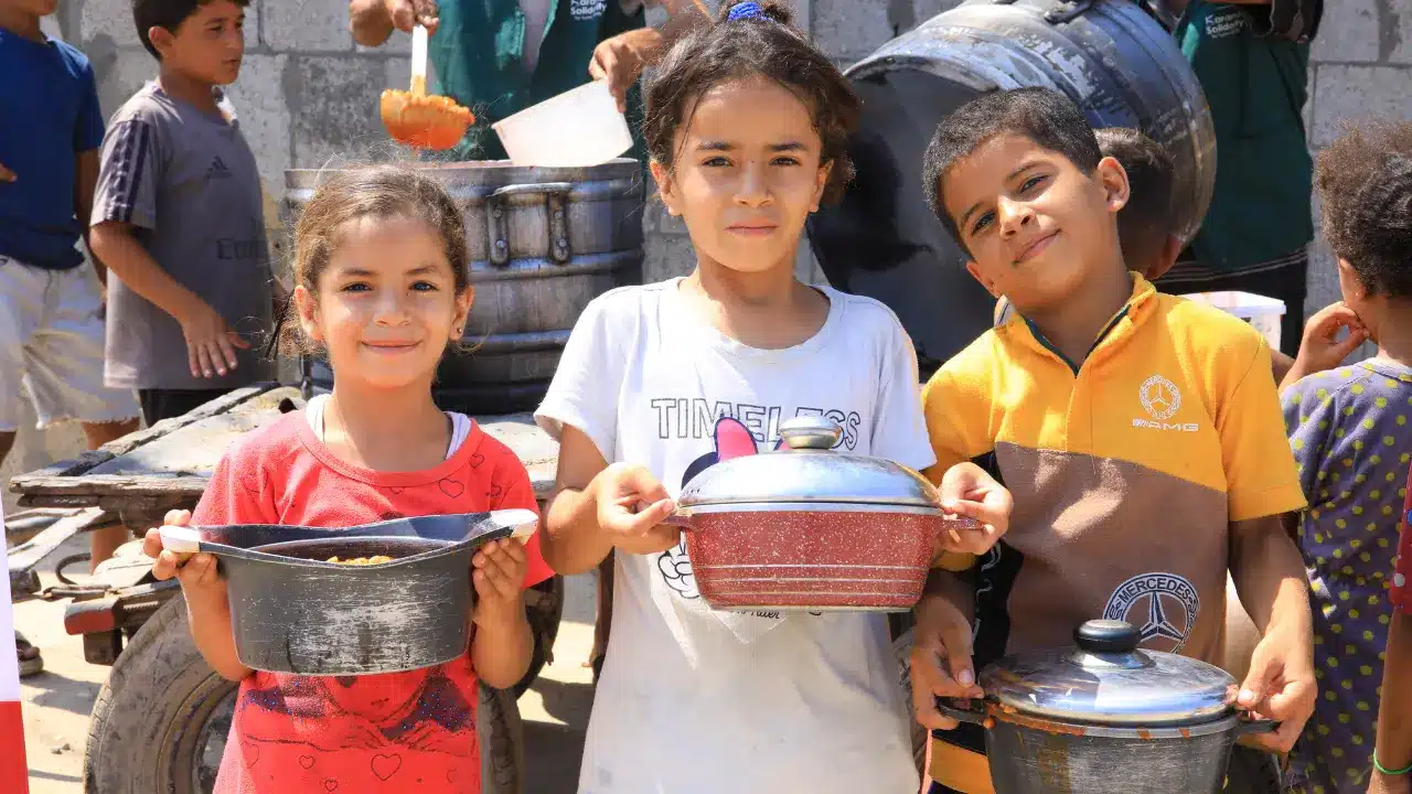 Trois enfants palestiniens souriant, tenant des casseroles pleines de repas nourrissants, en arrière-plan des bénévoles servant des repas chauds.