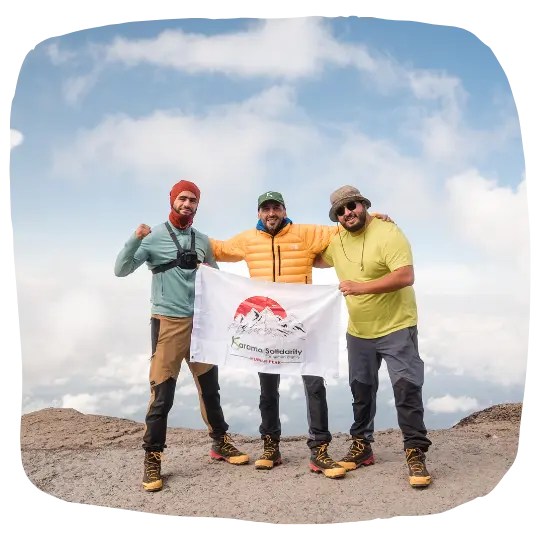 Trois hommes en tenue d'alpinisme, tenant une bannière Karama Solidarity au sommet d'une montagne, sous un ciel dégagé.