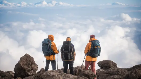 Trois randonneurs, équipés de sacs à dos et de bâtons de marche, contemplent un panorama époustouflant de nuages au sommet d'une montagne rocheuse.