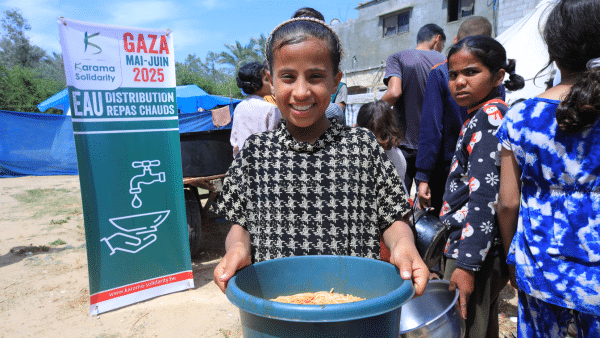 Une jeune fille palestinienne souriante tient un plat de pâtes, devant une file d’enfants et une banderole de Karama Solidarity annonçant la distribution de repas chauds à Gaza en mai-juin 2025.
